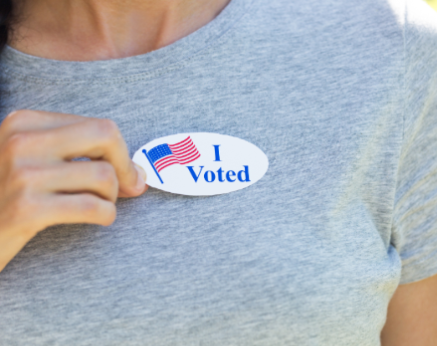 Woman wearing gray shirt and placing an oval sticker with I Voted in bold blue letters.
